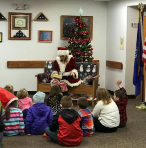 librarian dressed as Mrs. Clause reads to children in front of a Christmas tree
