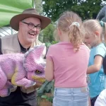 a man in a safari hat is smiling and holding a pink and yellow dinosaur in his arms as children feed it