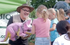 a man in a safari hat is smiling and holding a pink and yellow dinosaur in his arms as children feed it