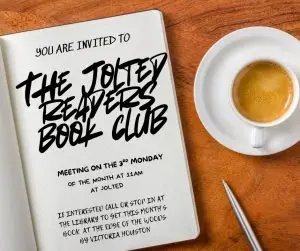 an open page of a book says "You are invited to The Jolted Readers Book Club. Meeting on the 3rd Monday of the month at 11AM." There is a cup of cappuchino and a pen next to the book
